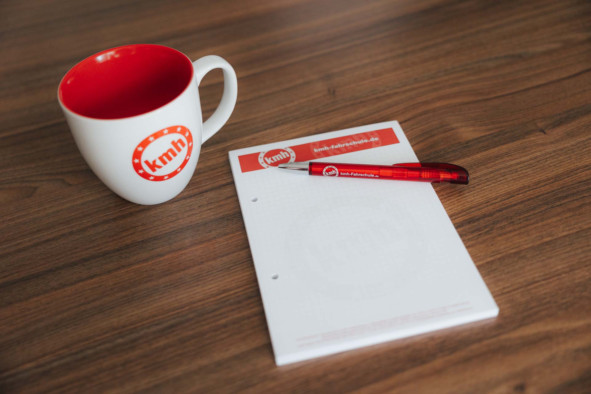 Kaffeetasse und Schreibblock mit Kugelschreiber. Alles in unserem Branding mit kmh Logo.